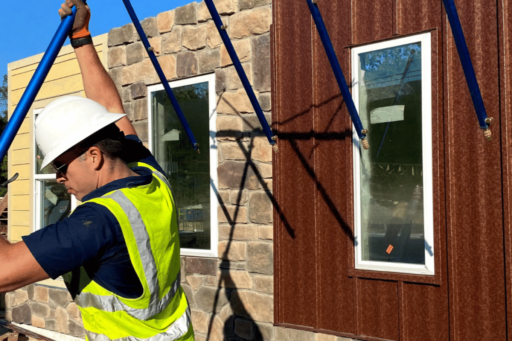 A man in a hard hat carries equipment near a window, preparing for a spray rack test.